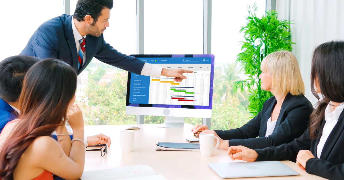 A man in a suit points at a computer monitor displaying a bar chart as four colleagues listen during a meeting in a bright office, discussing results from their HVAC business software.