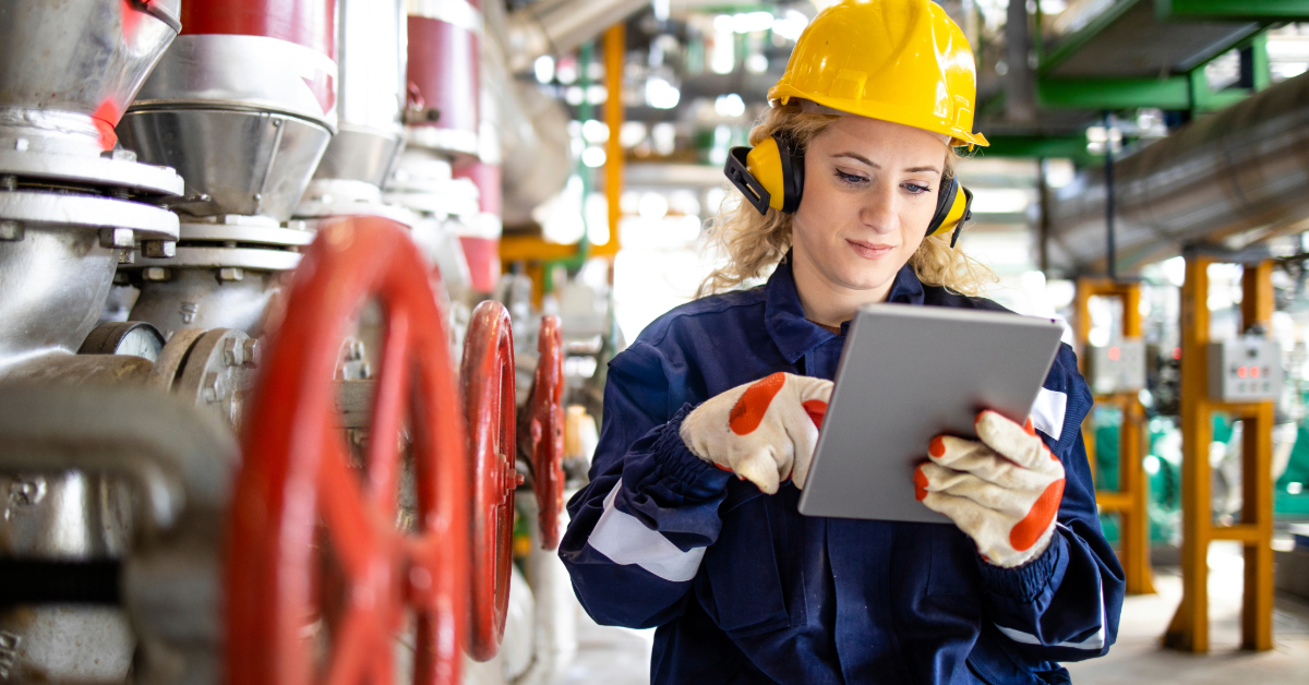 Woman in safety gear with a yellow helmet and gloves uses a tablet while standing in an oil and gas industry facility with pipes and large red valves, highlighting digital transformation on-site.