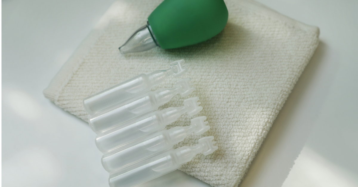 A green bulb syringe, often used as baby nasal aspirators, a stack of white ampoules, and a textured washcloth are arranged on a white surface.