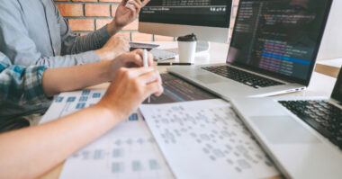 Two people work at a desk with laptops, a tablet, documents with charts, and code displayed on their screens as they follow a step-by-step guide for effective website localization.
