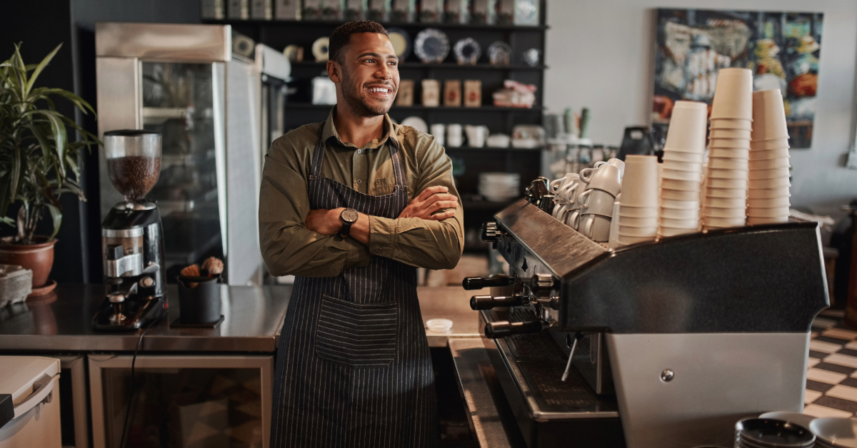 A barista wearing an apron stands with arms crossed and smiles behind the counter of a coffee shop, surrounded by pour-over coffee equipment and cups—a welcoming scene for aspiring coffee entrepreneurs.