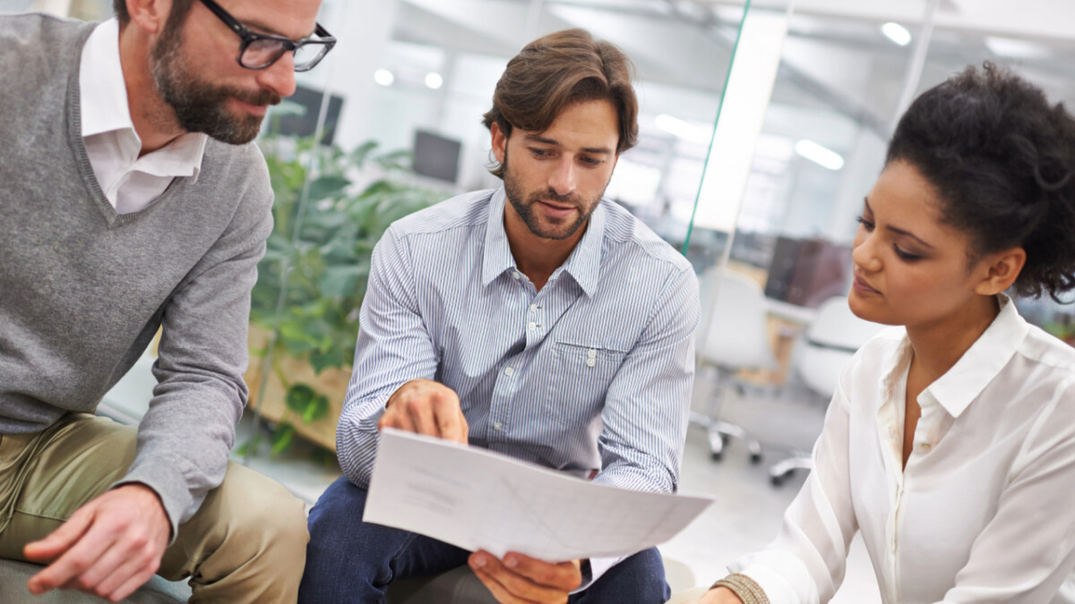 Three people sit together in an office, reviewing a document on startup funding trends. One person points at the paper while the others listen attentively, discussing projections for Q2 2024 and the future of their venture.