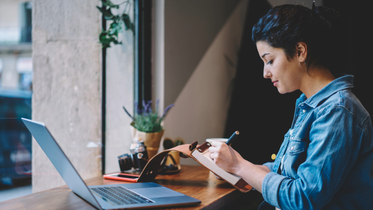 A woman sits at a window-side table working on a notebook with a pencil, creating helpful content to enhance reader experience, with a laptop, camera, and phone on the desk beside her.