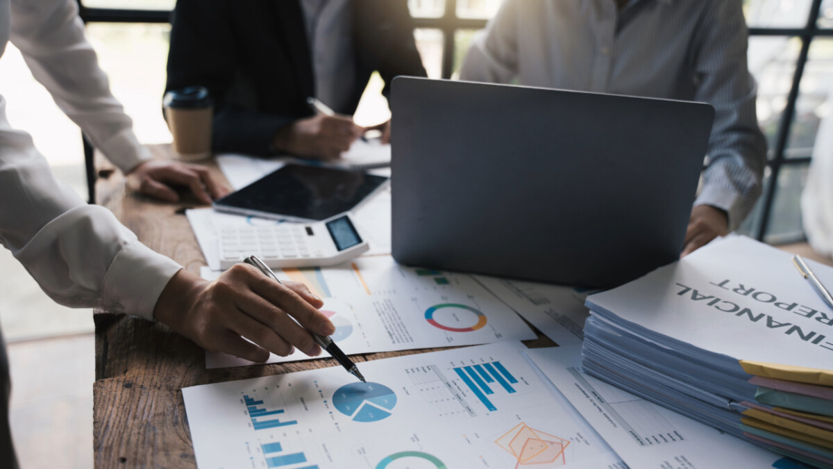 Three people sit at a desk with a laptop, calculator, coffee cup, and stacks of documents, reviewing printed financial reports and charts as they discuss the importance of business investment for future growth.