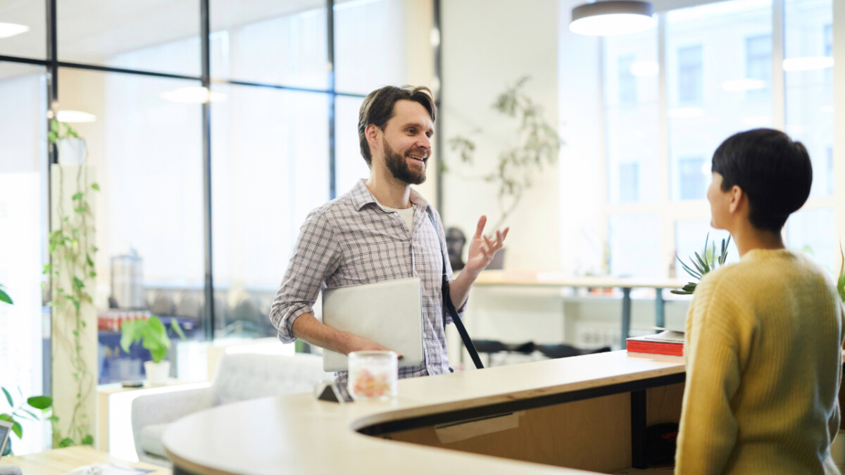 A man holding a laptop stands at a reception desk, discussing the hidden aspects of running a hospitality business with a seated woman in a modern office setting.
