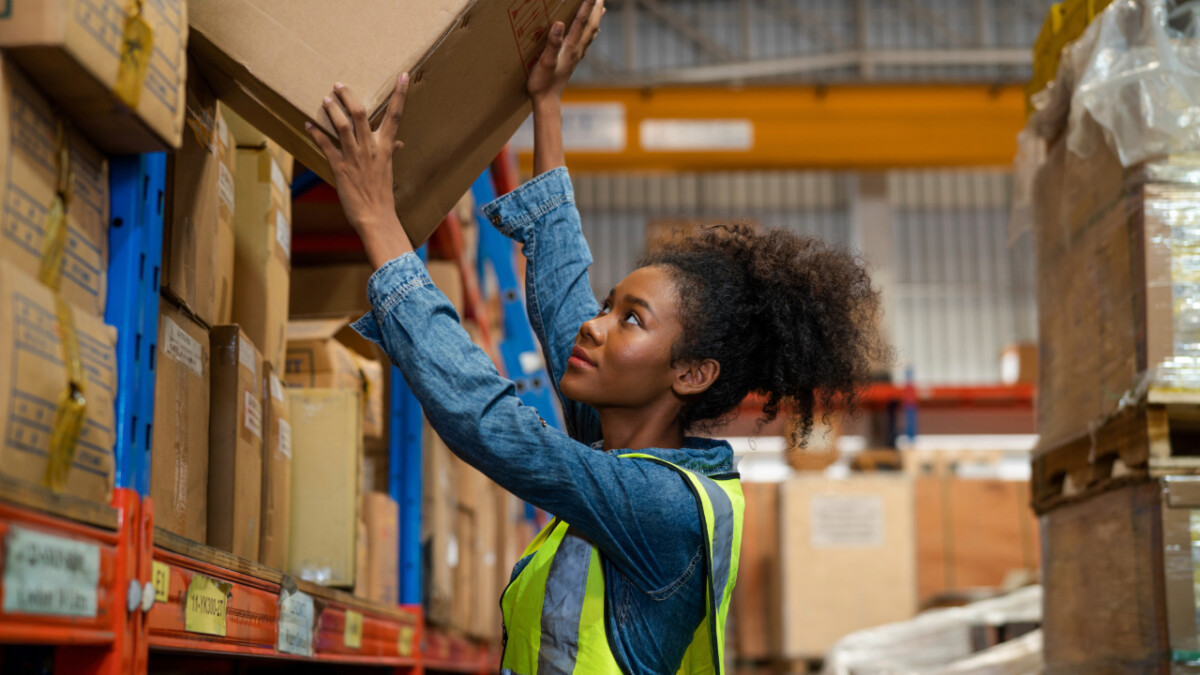 A woman wearing a reflective vest reaches up to retrieve a cardboard box from a warehouse shelf, demonstrating proper warehouse storage safety.