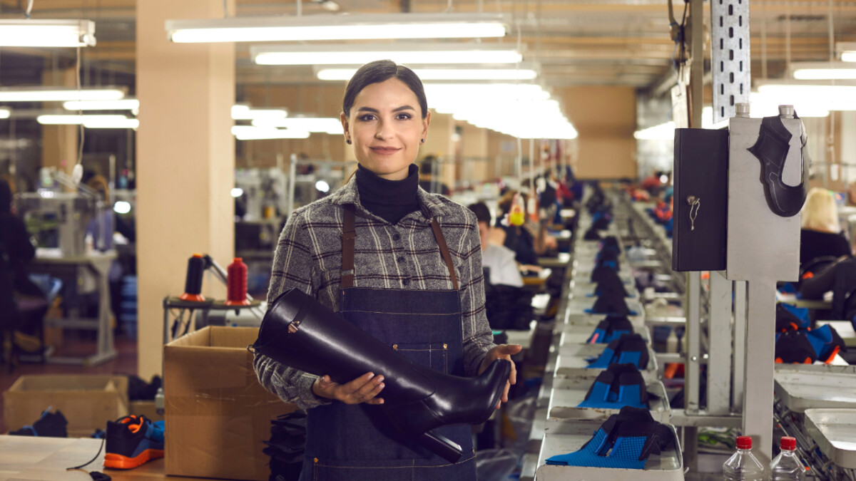 A woman in a factory holds a black boot while standing by a workbench, offering production tips as other workers and assembly lines mass-producing footwear are visible in the background.