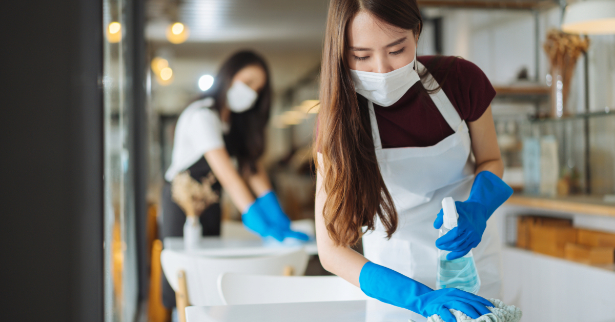 Two women wearing masks and gloves clean tables in a restaurant, one in the foreground wiping with a spray bottle of food-grade cleaning products, the other cleaning in the background.