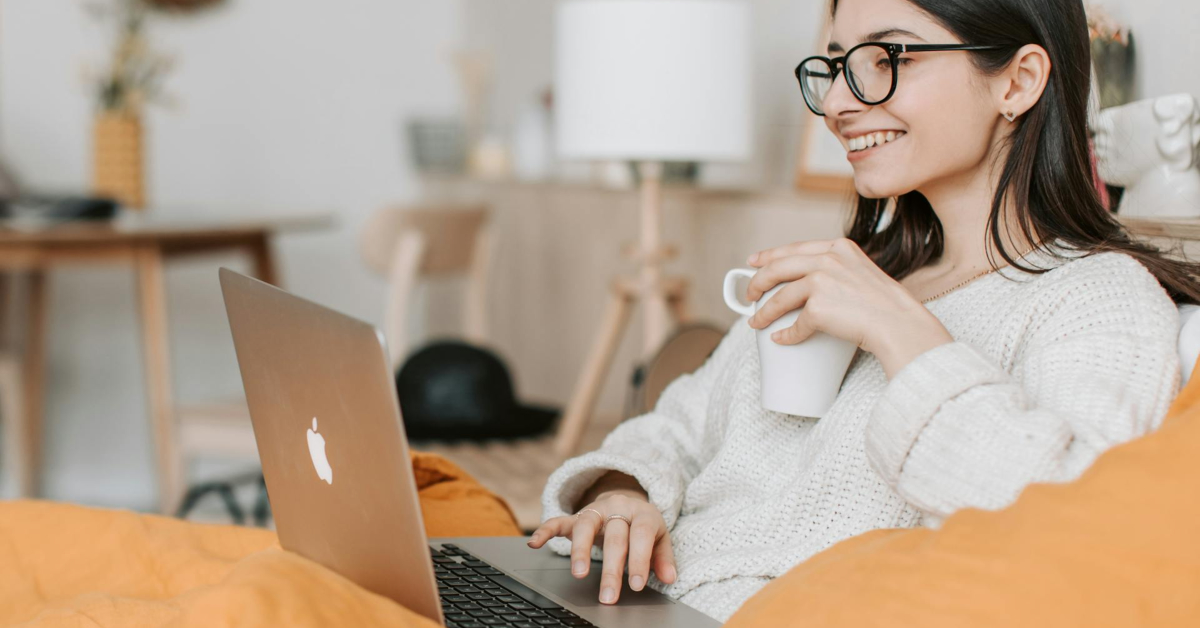 A woman wearing glasses sits on a bed with a yellow blanket, smiling while using her laptop to discover ecommerce tips, holding a white mug in her other hand.