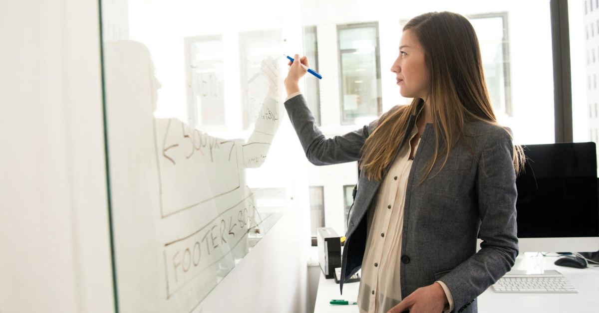 A woman in business attire writes diagrams on a glass board in a modern office, analyzing reasons business fails with colleagues as large windows and computer monitors surround them.