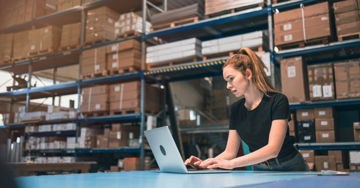 A woman using a laptop at a workstation in a warehouse with shelves of boxes in the background, managing E-Commerce Manufacturing operations and ensuring UFLPA Compliance.