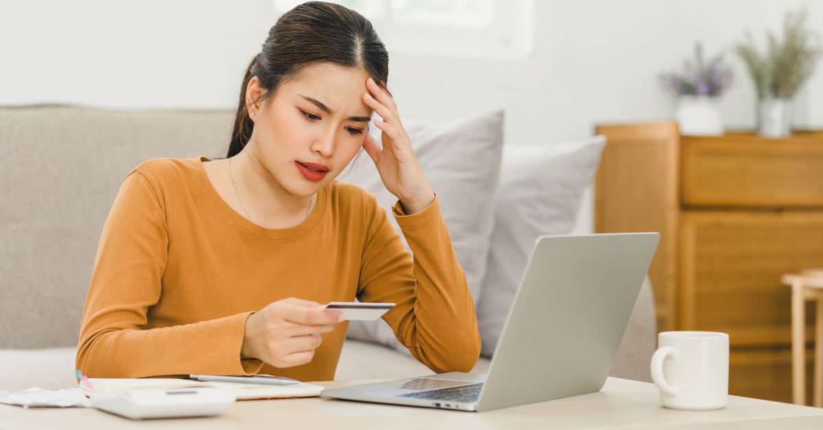 A woman sits at a table looking concerned while holding a credit card and staring at a laptop, worried about the chargeback impact on merchants; a notebook, calculator, and coffee mug are nearby.