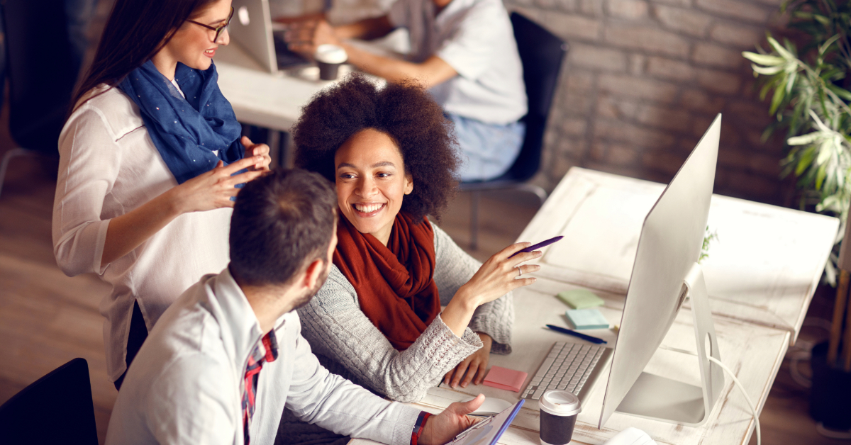 Three people are having a discussion at a shared desk in an office setting, with computers, notebooks, and coffee cups visible as they explore modern solutions to propel your business forward.