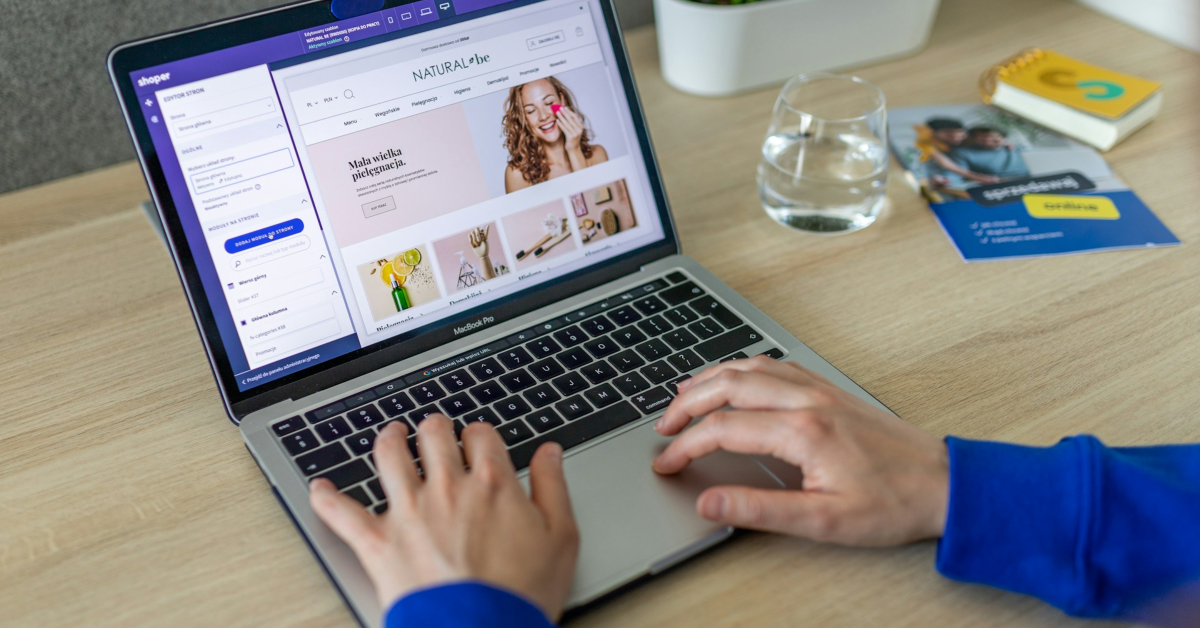Person using a laptop to design an ecommerce website at a desk, with a glass of water, plant, and notebook nearby—perfecting the online store’s look while considering ecommerce SEO for better visibility.