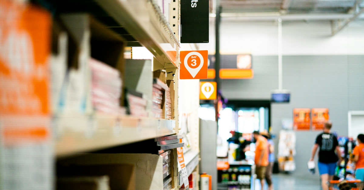 A hardware store aisle with shelves of business supplies, an orange aisle 3 sign, and several people browsing in the background near the entrance, choosing materials for their next project.