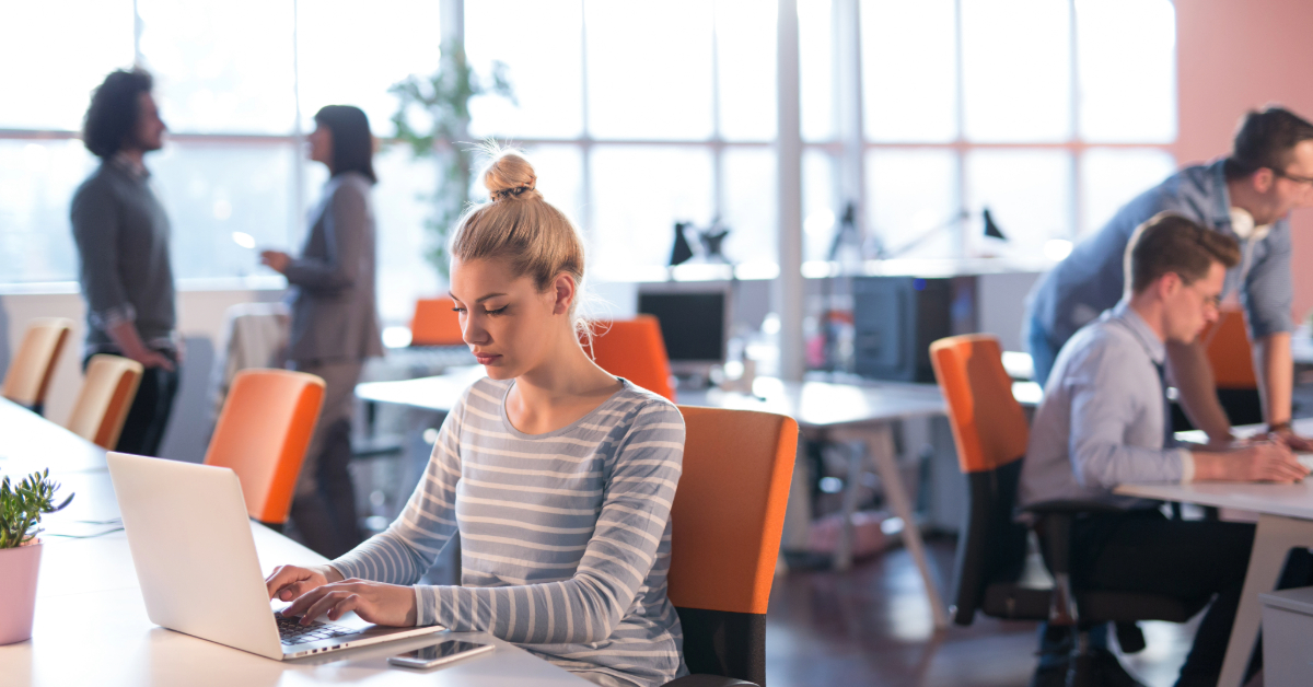 Woman works on a laptop at a desk in a modern open-plan office with excellent office amenities, while other people collaborate and talk in the background.