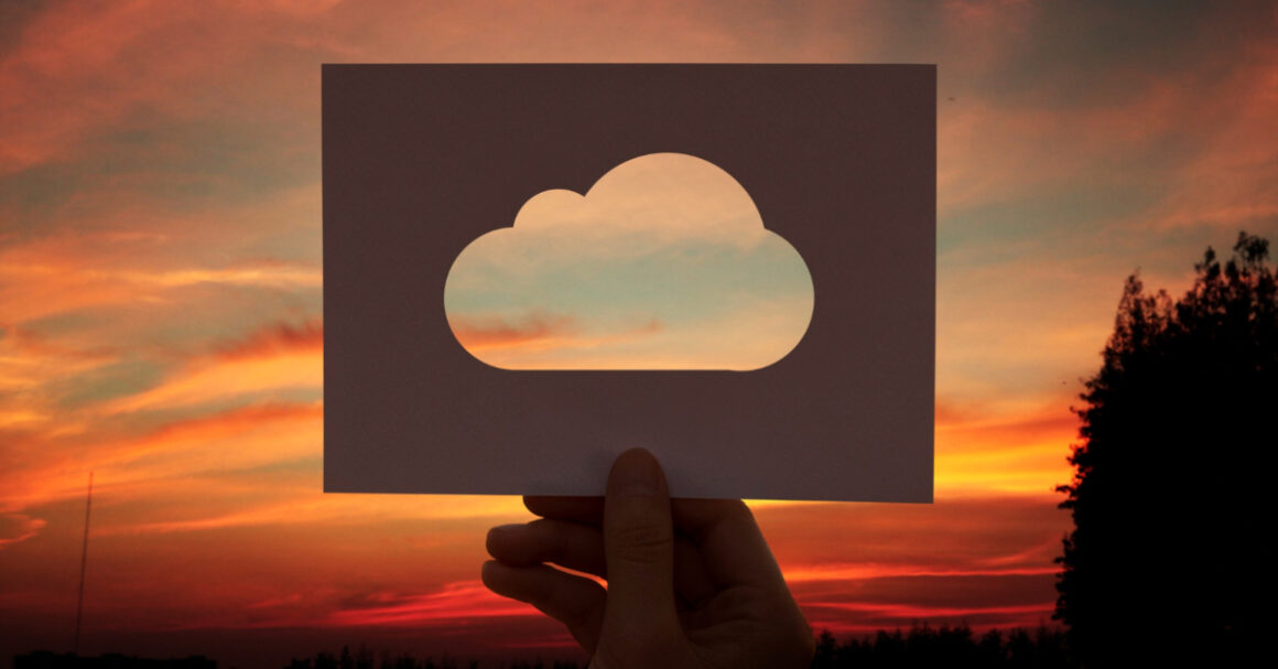 A hand holds a piece of paper with a cloud-shaped cutout against a colorful sunset sky, symbolizing innovative Cloud Solution Development by Euristiq’s IT experts.