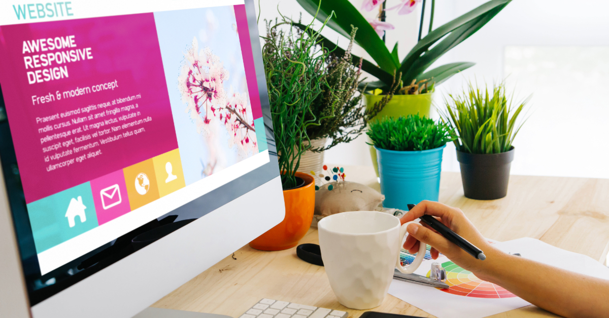 A web designer works at a desk with a computer displaying a web design, surrounded by plants, a notebook, and a coffee mug.