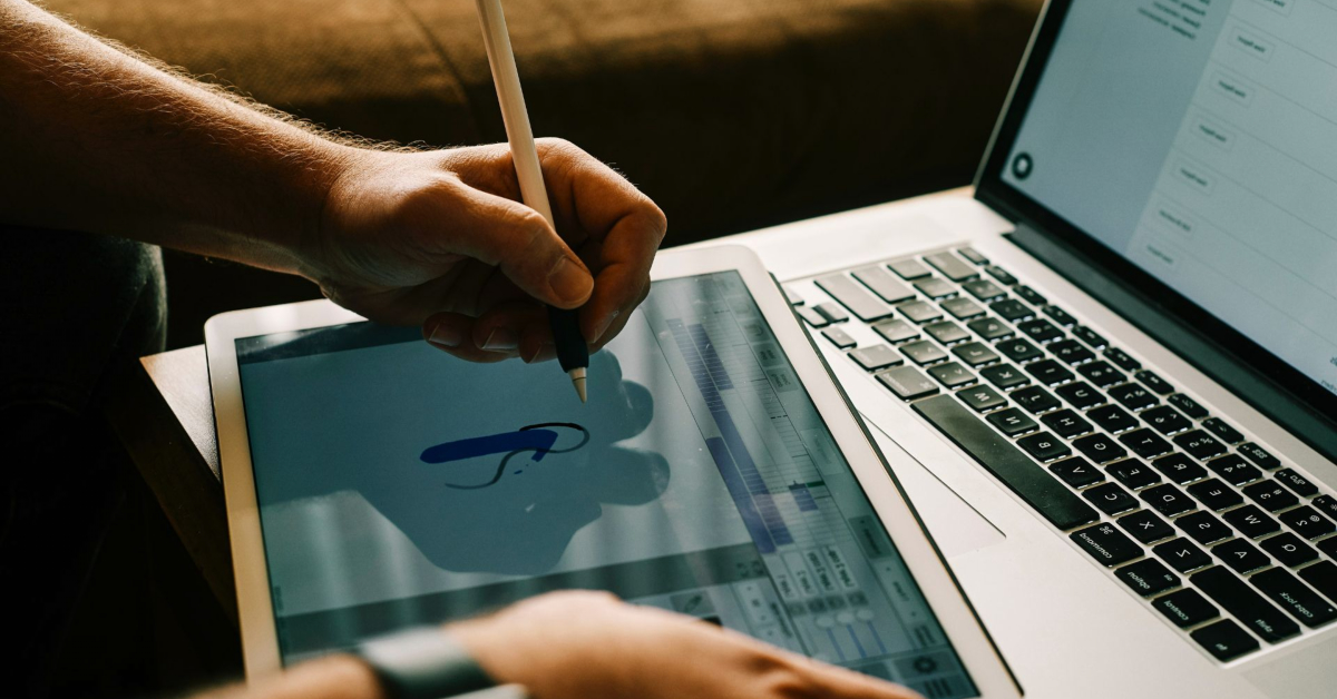 A person uses a stylus to draw on a tablet with graphic design software while a laptop displaying a spreadsheet is open beside them, showcasing modern design tools in 2024.