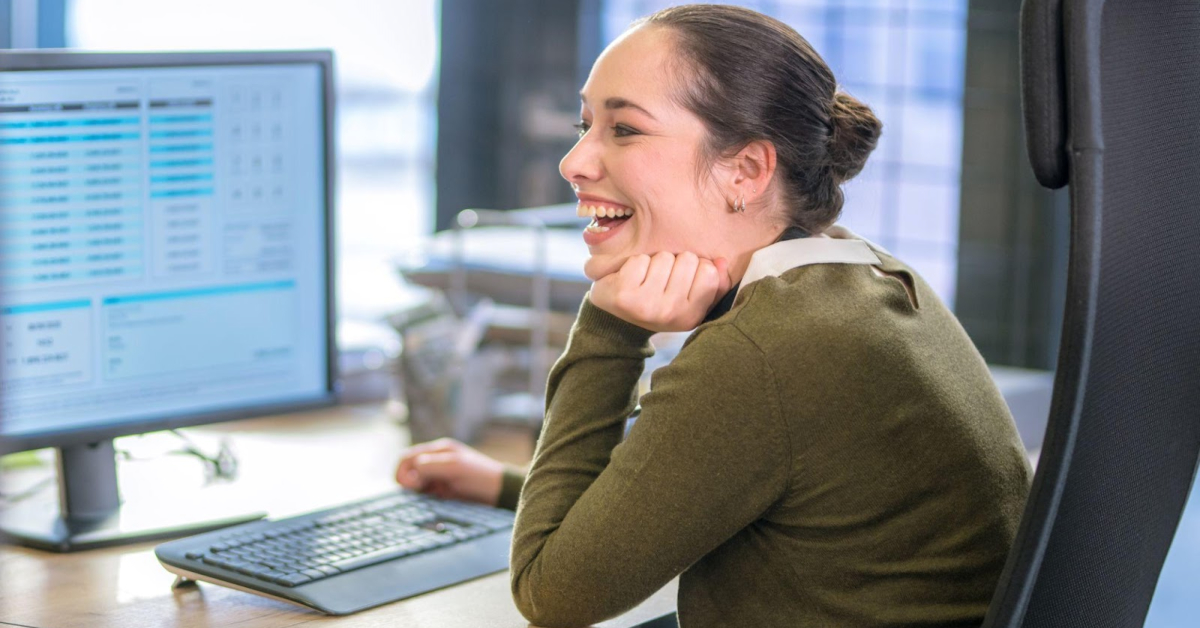 A woman sits at a desk, smiling while looking at a computer monitor with spreadsheets or CX data displayed on the screen, highlighting the efficiency of Ecommerce Outsourcing in Costa Rica.