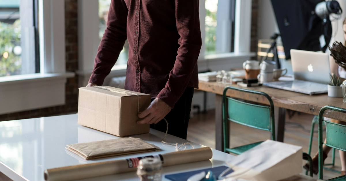 A person stands at a table tying string around a cardboard box, with envelopes, paper, and office materials nearby, showcasing the careful packing process typical in order fulfillment for modern businesses.