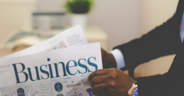 A person in a suit is holding and reading a business newspaper at a desk, surrounded by business essentials—perfect for anyone starting a new business—with a blurred plant in the background.