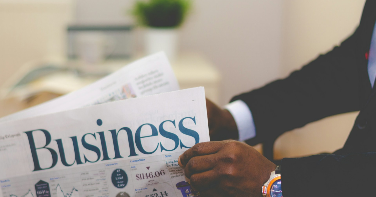 A person in a suit is holding and reading a business newspaper at a desk, surrounded by business essentials—perfect for anyone starting a new business—with a blurred plant in the background.
