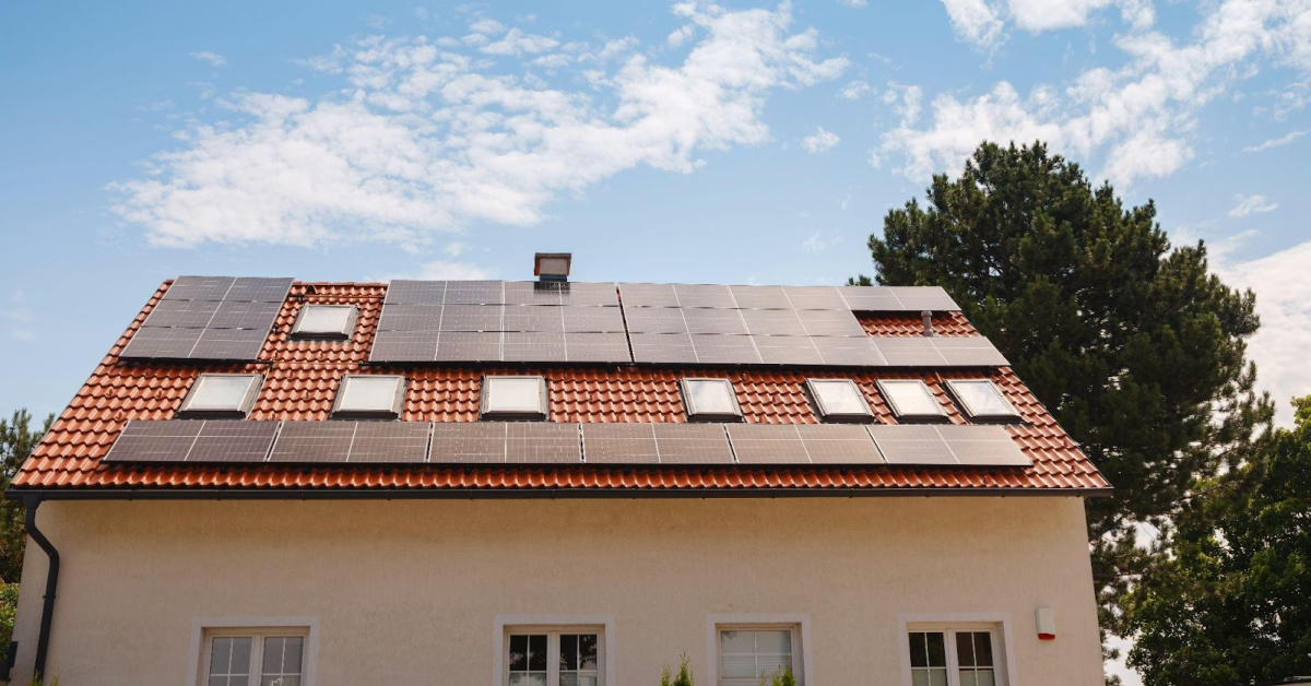 A house with a red tile roof covered in solar panels, set against a blue sky and scattered clouds, offers both beauty and the chance to save money through long-term financial savings. Trees complete the peaceful background.
