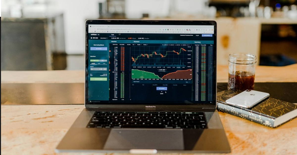 A laptop displaying a financial trading platform with currency markets data sits on a wooden table beside a drink, a notebook, and a smartphone, highlighting the convenience of online trading with c-markets-co.com.