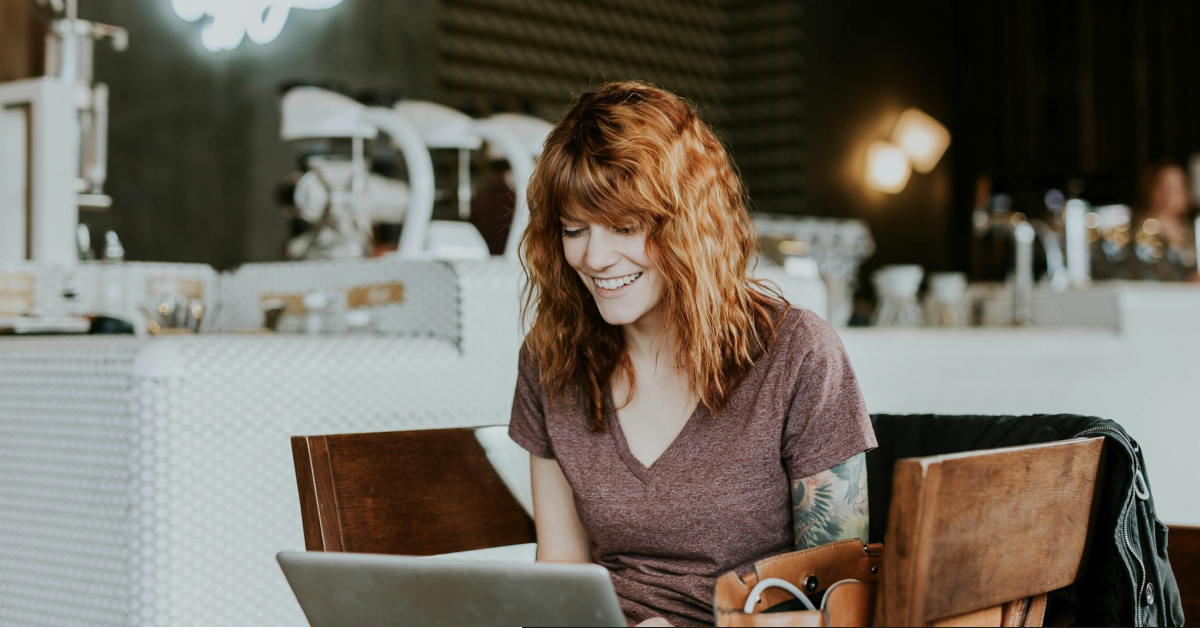 A woman with wavy red hair sits at a café table, smiling as she looks at her open laptop, exploring online side hustles to make money.