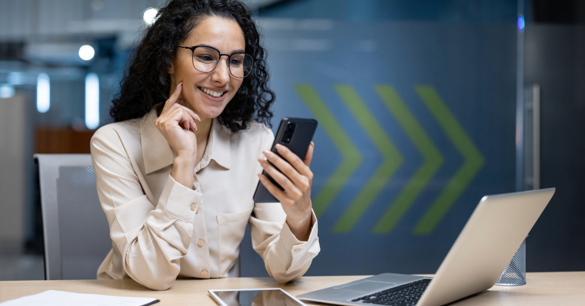 A woman wearing glasses sits at a desk with a laptop and tablet, smiling while looking at her smartphone, possibly reading a guide on VoIP texting in a modern office setting.