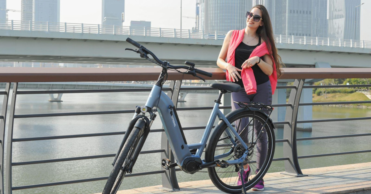 A woman in sunglasses stands by a railing next to a light blue electric bicycle, highlighting the rising popularity in the electric bicycle market, with a river and city buildings visible in the background.