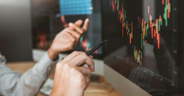 Two people discuss and point at computer monitors displaying colorful stock market charts with candlestick patterns as they learn pin bar strategies for better trading decisions.