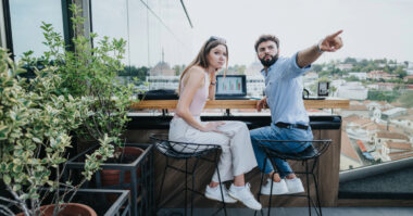Two people sit at a rooftop table with a laptop displaying graphs; the man points into the distance while the woman looks at the camera—capturing a moment from their beginner's guide on how to turn passion into profit.