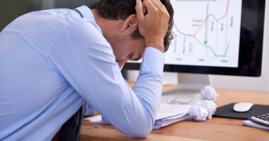 A man in a dress shirt sits at a desk with his head in his hands, surrounded by papers and crumpled notes, searching for ways to banish burnout as a declining line graph is displayed on his computer screen.