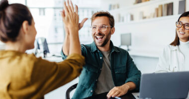 Three people in an office celebrate a success; one man smiles and gives a high-five to a woman while another woman watches and smiles, demonstrating how effective networking can boost your career, with a laptop visible on the table.
