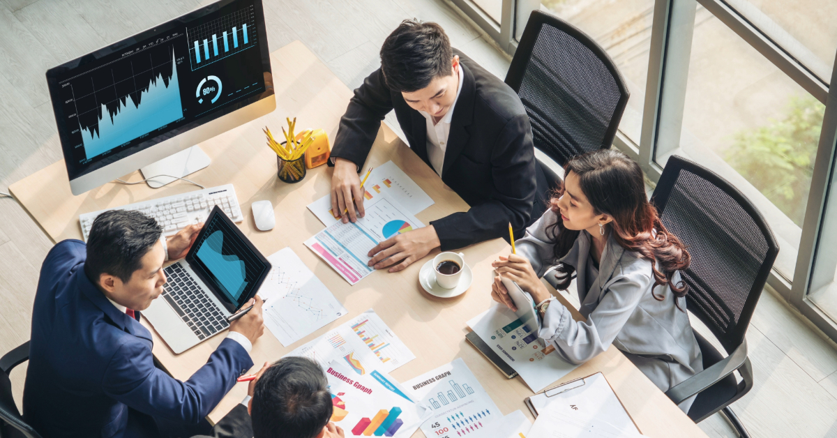 Four businesspeople sit around a table with charts, graphs, and laptops, discussing a content audit. A monitor displays financial graphs as sunlight enters through large windows.