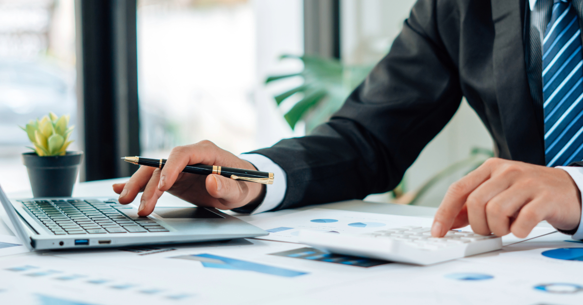 A person in a suit uses a laptop and a calculator at a desk covered with charts and papers, carefully focusing on tax planning.