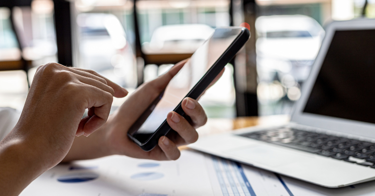 A person uses a smartphone next to an open laptop on a desk with papers and charts, ensuring data accuracy while reviewing phone number best practices.