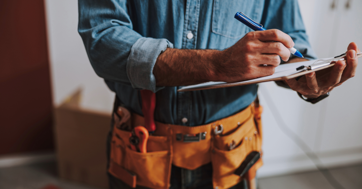 A person wearing a tool belt and denim shirt, equipped with the necessary skills, writes on a clipboard with a blue pen.