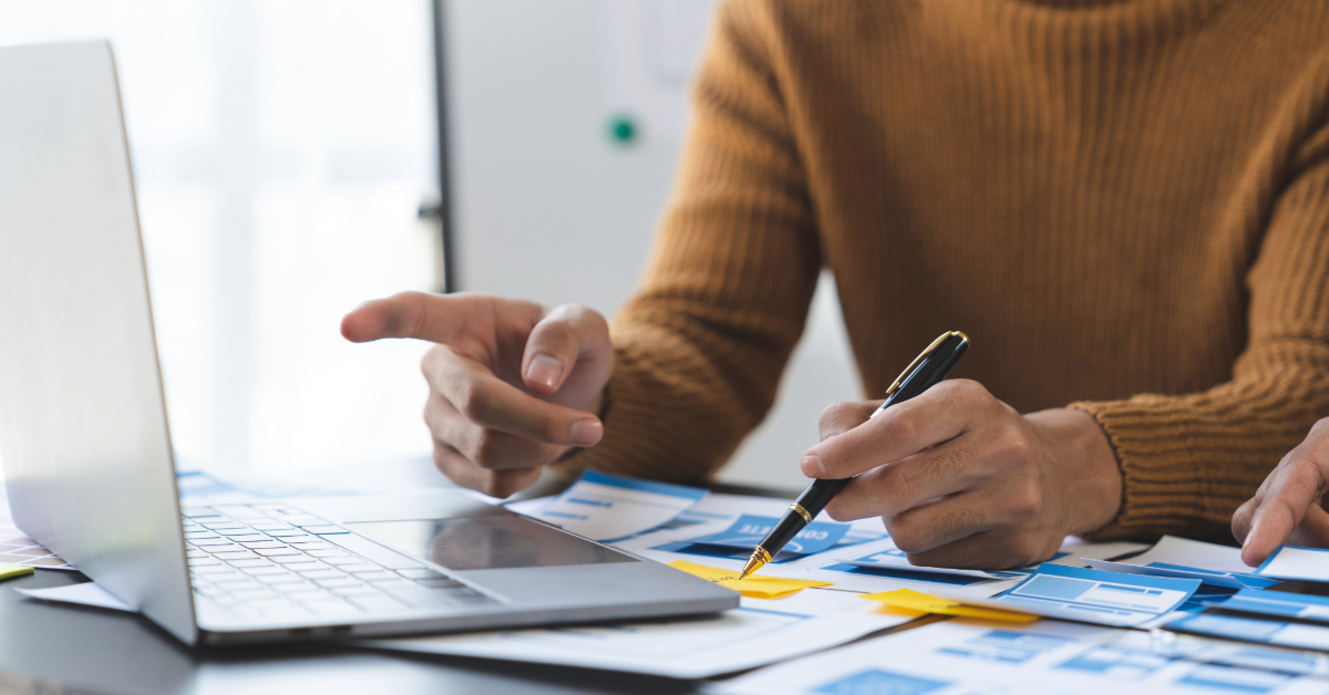Two people work together at a desk with a laptop and documents, discussing plans and pointing at papers during a meeting, collaborating on Shopify development services.