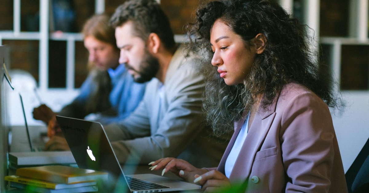 Three people sit in a row at desks, working on laptops in a modern office setting. The focus is on a woman in the foreground wearing a light purple blazer, efficiently managing multiple domain names with ease.