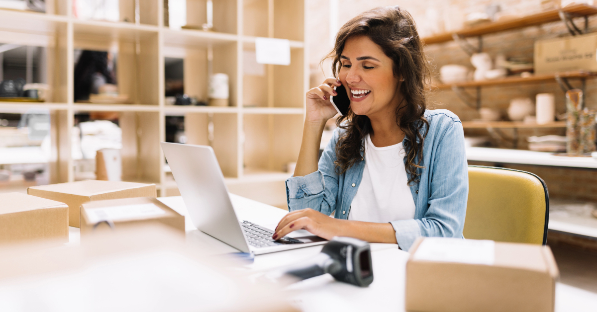 A business owner sits at a desk with her laptop, talking on the phone, surrounded by cardboard boxes in an eCommerce workspace or small business setting.