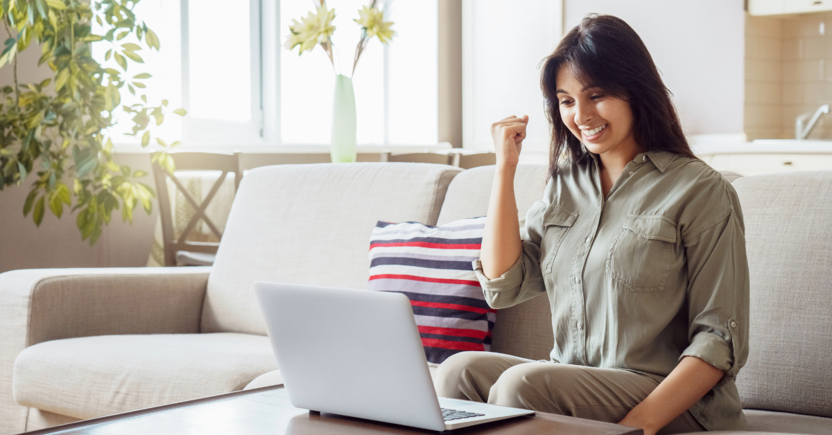 A woman sits on a sofa in a living room, smiling and raising her fist in excitement while looking at a laptop—celebrating a big win with her favorite unique eCommerce brand.