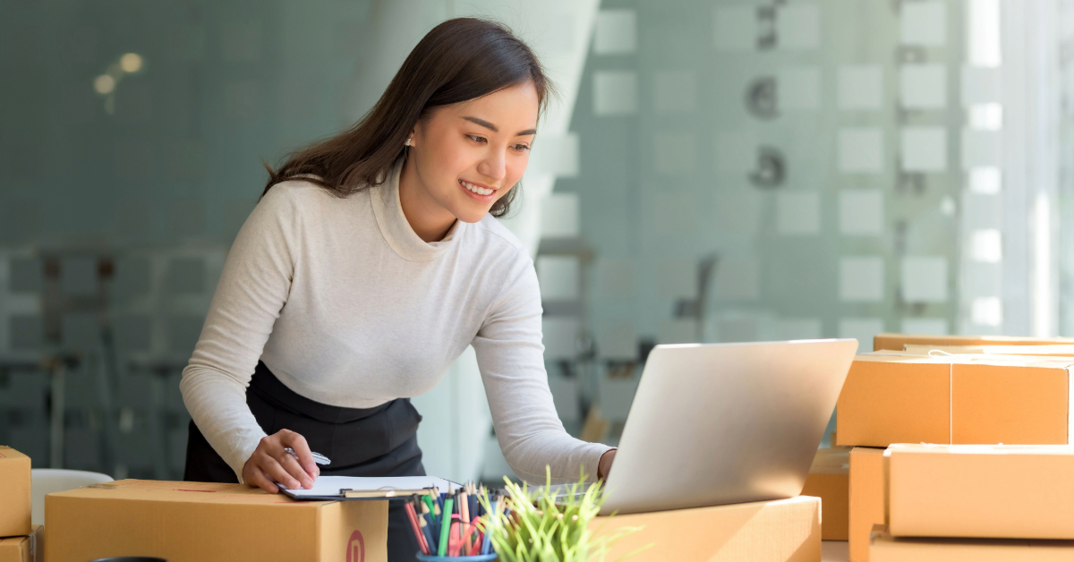 A woman stands at a desk, looking at a laptop and holding a clipboard, surrounded by cardboard boxes—planning marketing strategies for her online business in an office setting.