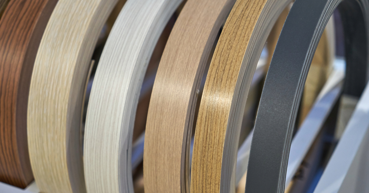 Several rolls of edge banding in various colors and wood grain patterns are displayed vertically on a rack, showcasing the versatile selection available from Framatech.