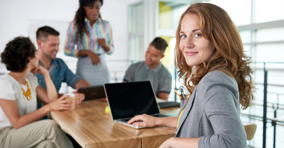 A woman sits at a desk with a laptop, looking at the camera, while four colleagues discuss high-performance agency tactics in the background.