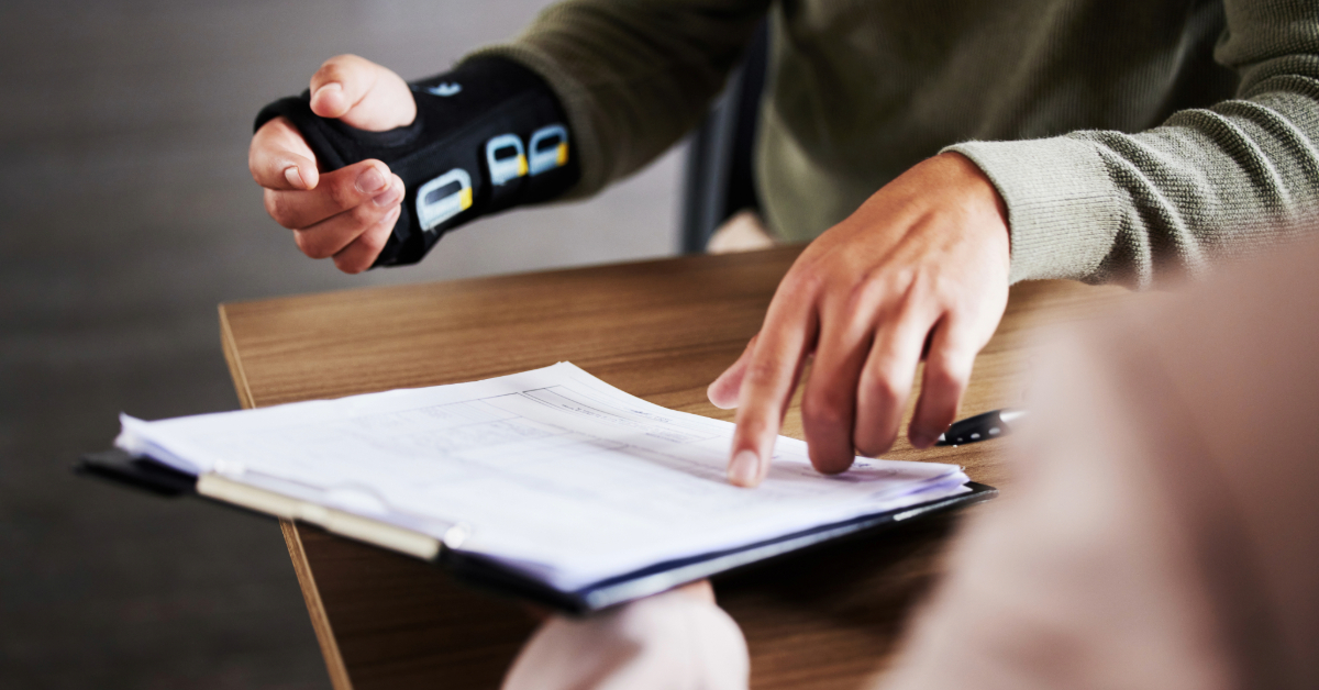 A person wearing a wrist brace, possibly due to a work injury, holds a pen and gestures toward documents on a clipboard, while another person sits across the table.