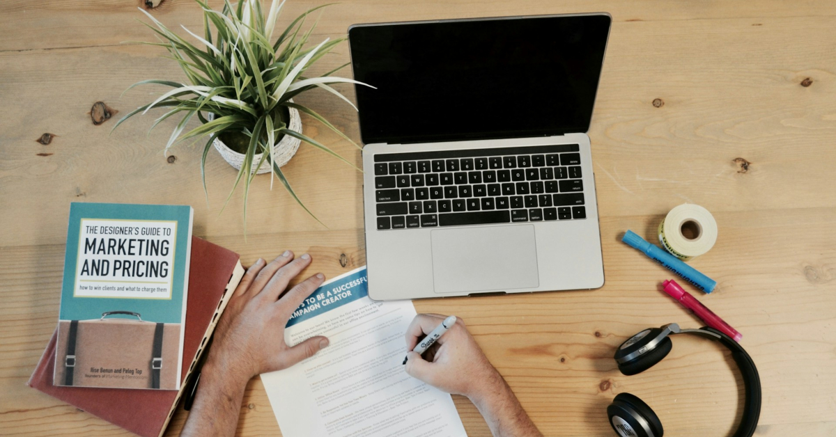 A person writes on a form at a wooden desk with a laptop, plant, books, headphones, tape, and pens—showcasing the organized workspace of digital marketing assistants driving ecommerce growth.