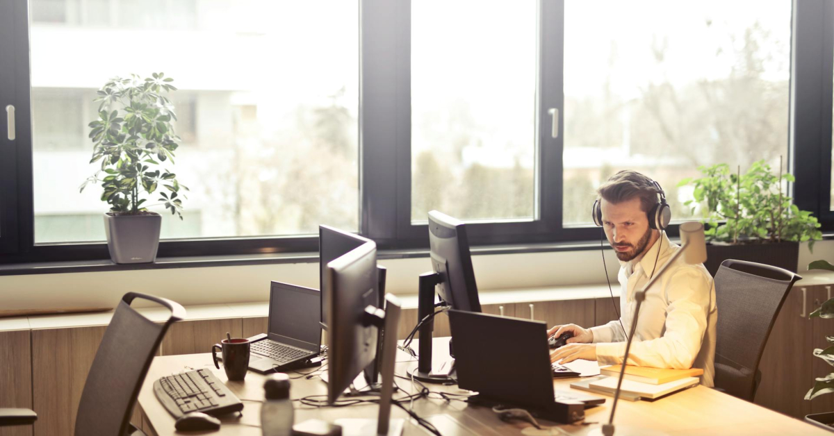 Man wearing headphones sits at a desk with multiple computer monitors, ensuring business transactions remain secure in a bright office with large windows and potted plants.